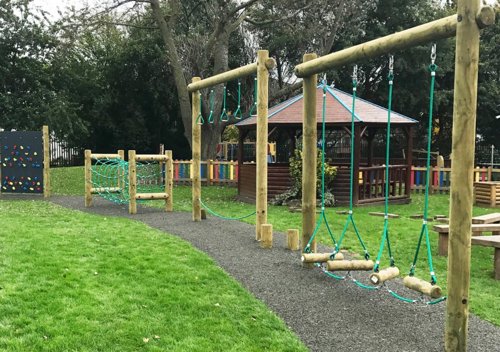 Timber trim-trail playground with hanging rope steps, monkey rings, and net tunnel beside a gazebo.