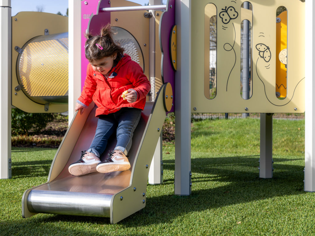 Toddler in a red jacket sliding down a small metal slide on a low playground structure.