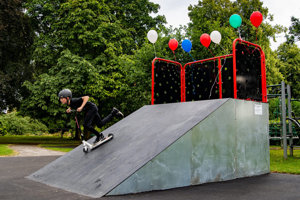 Teen riding a scooter up a metal skate ramp decorated with balloons in a park.