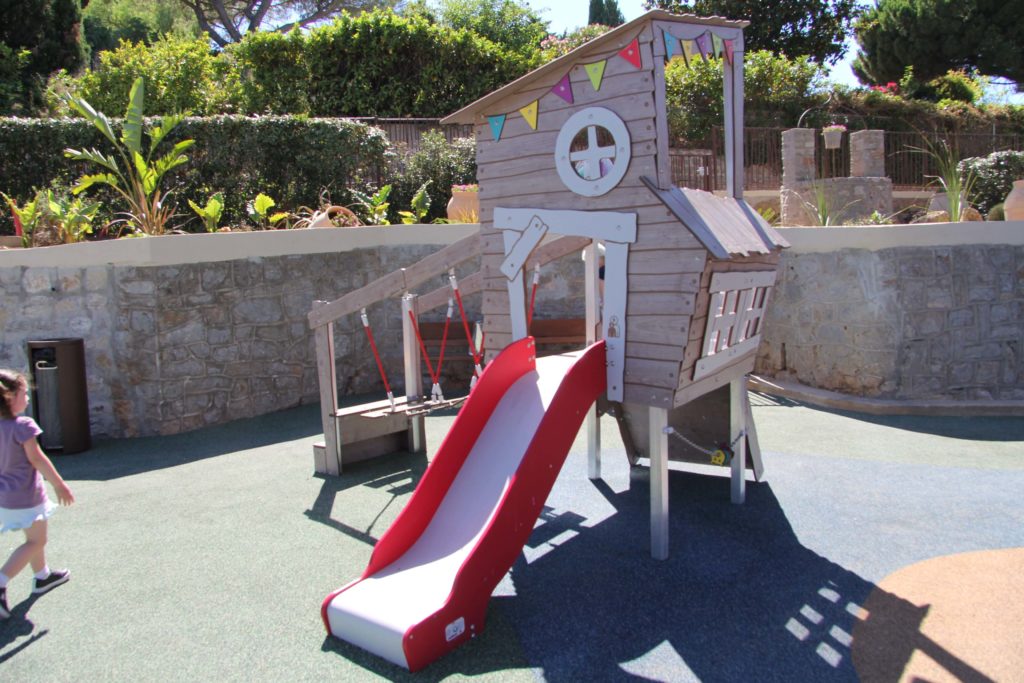 Small wooden playhouse structure with a red slide and rope bridge on rubber safety surfacing.