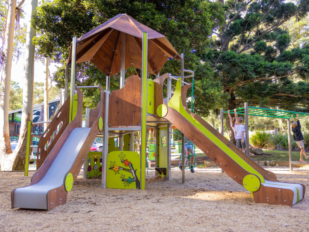 Large wooden playground tower with two slides and children playing nearby in a wooded park.