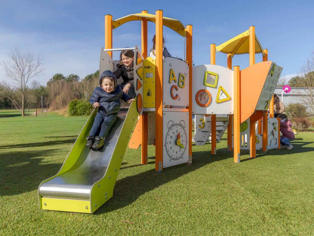 Toddlers playing on a compact orange-and-yellow playground with slide and activity panels on grass.