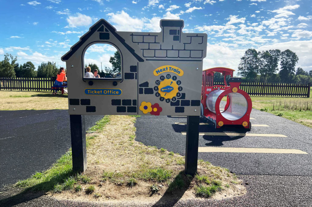 Playground role-play panel shaped like a ticket office beside a small red train-themed tunnel.