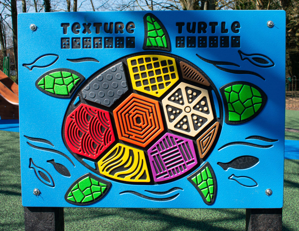 Bright blue sensory playground panel featuring a textured turtle design with raised patterns and braille.