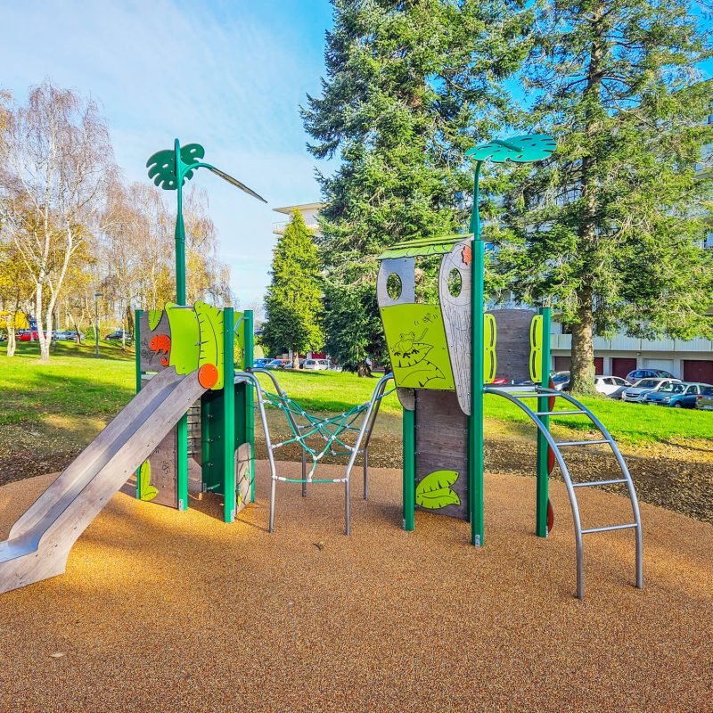 Green nature-themed playground unit with slide, climbing net, and arched ladder in a park.